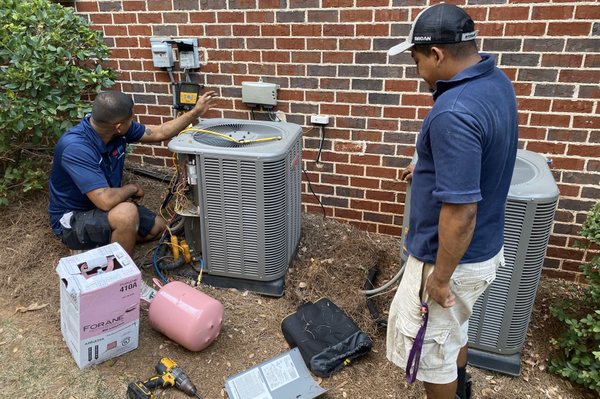 HVAC Guyz & Plumbing Inc — two technicians in branded blue shirts working on a residential AC condenser on brick-home side yard