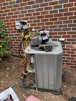 HVAC Guyz & Plumbing Inc — residential AC condenser with overhead disconnect box and refrigerant lines against a brick home